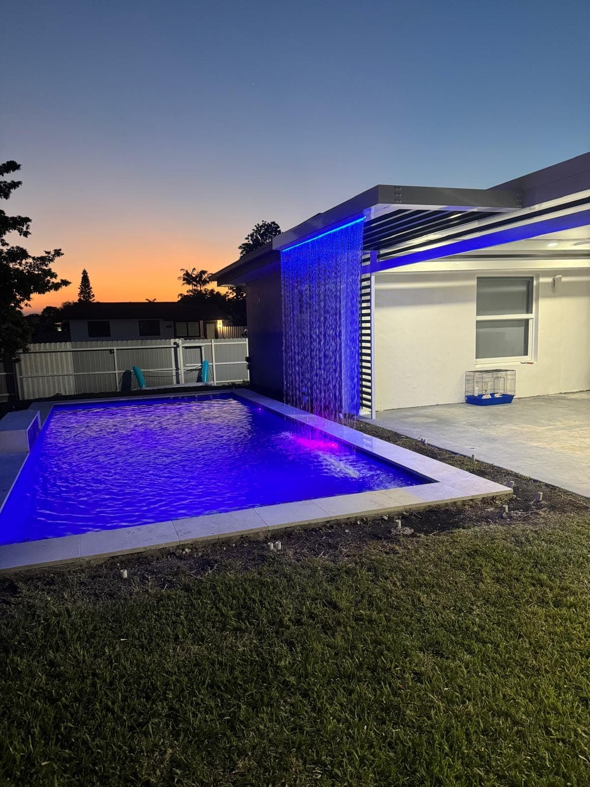 Pérgola de aluminio con cortina de agua y luces LED sobre la piscina al atardecer