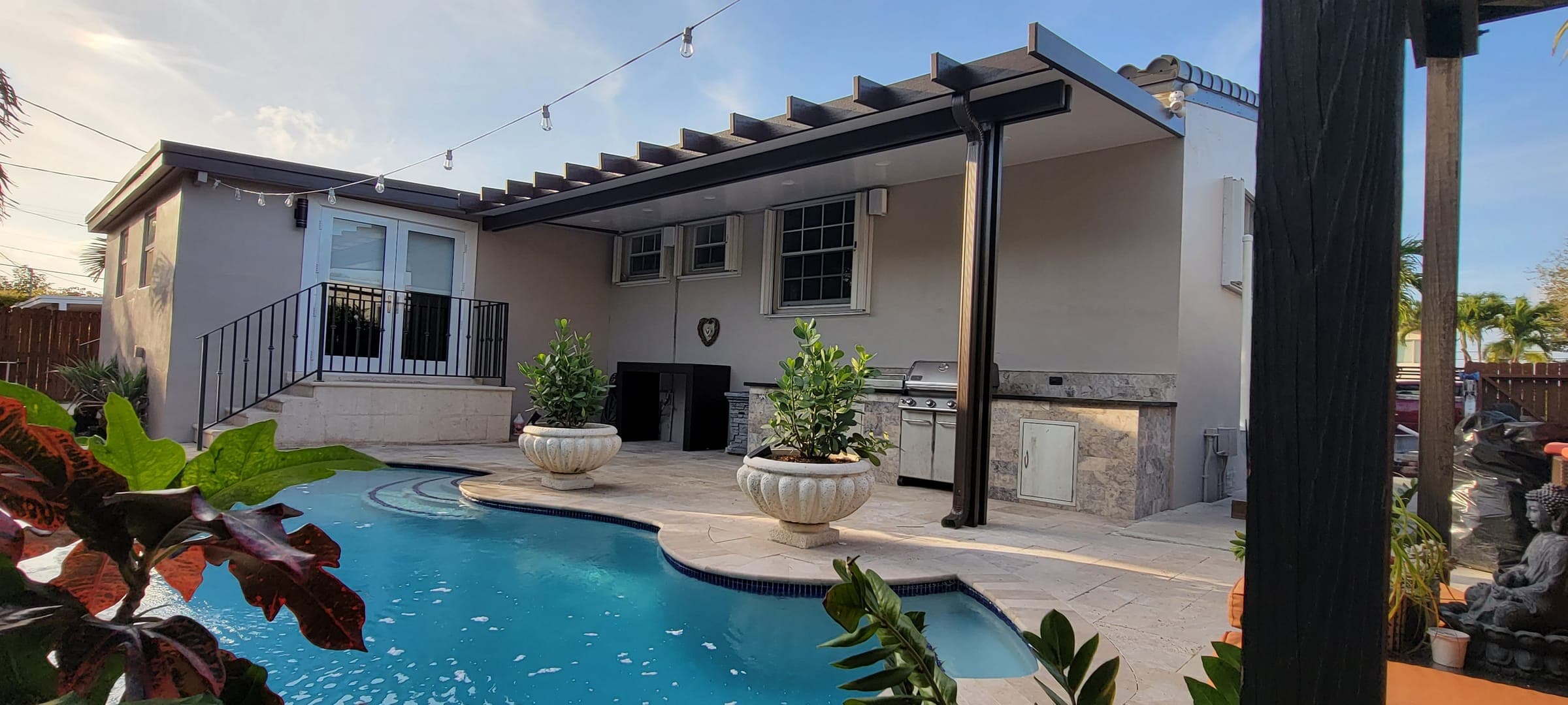 Cocina de exterior en piedra con parrilla bajo pérgola de aluminio bronce junto a la piscina